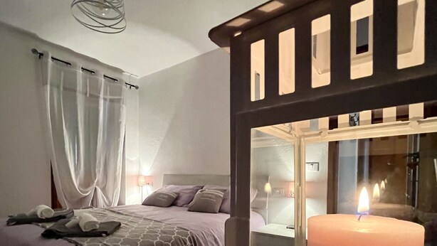 Une chambre à coucher avec un lit, une table de chevet, une commode, une fenêtre avec une bougie et un luminaire suspendu.