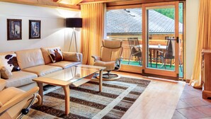 Ein modernes Wohnzimmer mit Sofa, Sessel und Glastisch. An der Wand hängen zwei gerahmte Bilder, ein großes Fenster mit Vorhängen und eine Glastür, die zu einem Außenbereich mit Tisch und Stühlen führt.