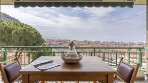 Ein Holztisch mit zwei Stühlen und einer dekorativen Vase, mit Blick auf eine Stadtlandschaft und eine Wasserfläche sowie einen Hügel.