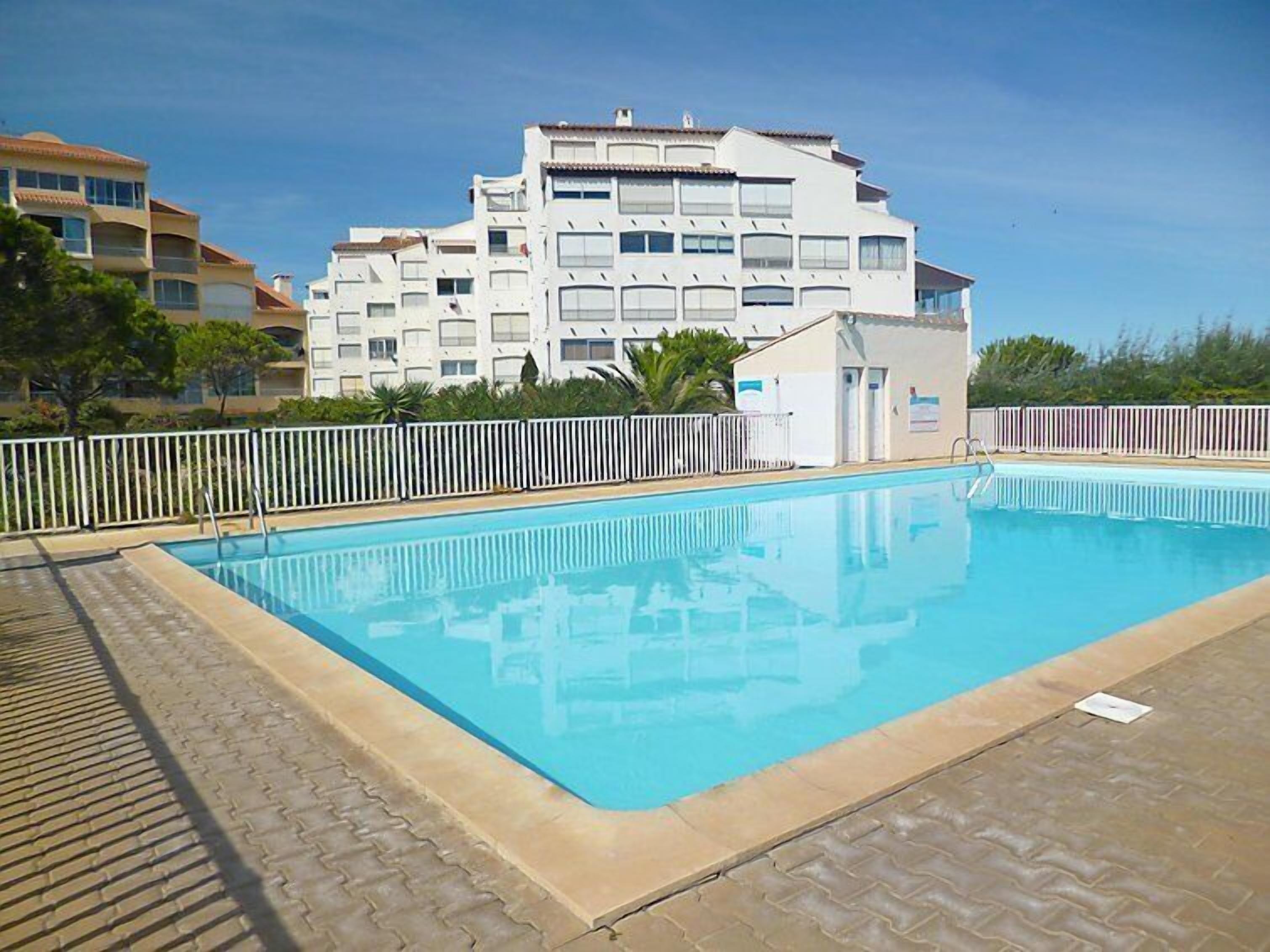 Una piscina rectangular con borde de azulejos, rodeada por una cerca blanca. Al fondo, se observa un edificio de varios pisos con balcones y techo plano.