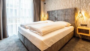 Habitación de hotel con una cama grande, una mesita de noche, una lámpara de pared y una ventana con cortinas.