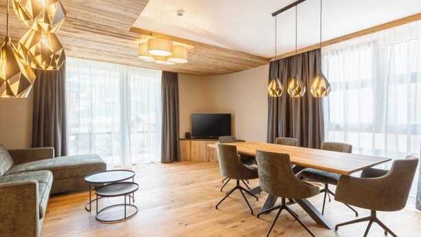 Una habitación de hotel moderna con techo de madera, una amplia mesa de comedor con sillas, un sofá, una pequeña mesa redonda y un televisor.