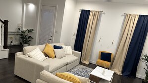 Una sala de estar con un sofá color beige, un sillón amarillo y una mesita con un jarrón blanco. Hay dos cortinas en las ventanas, una azul y otra beige.