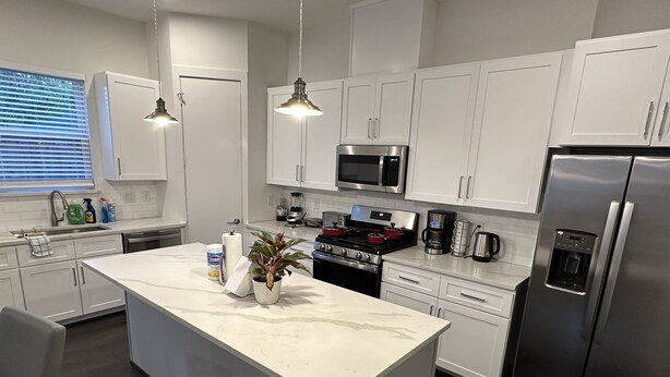Una cocina moderna con armarios blancos, electrodomésticos de acero inoxidable, una isla de mármol e iluminación colgante.