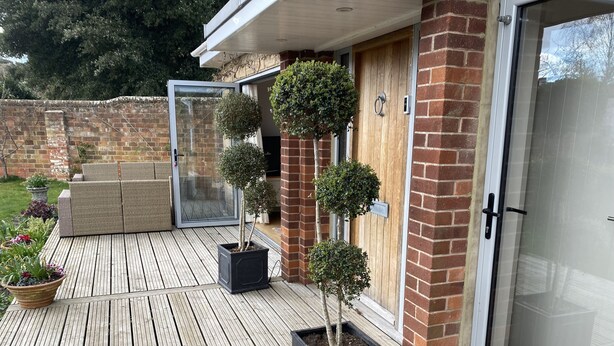 A wooden deck with potted plants, a wicker sofa, and a sliding glass door leading to a brick wall. - East Park Lodge (Cowes)