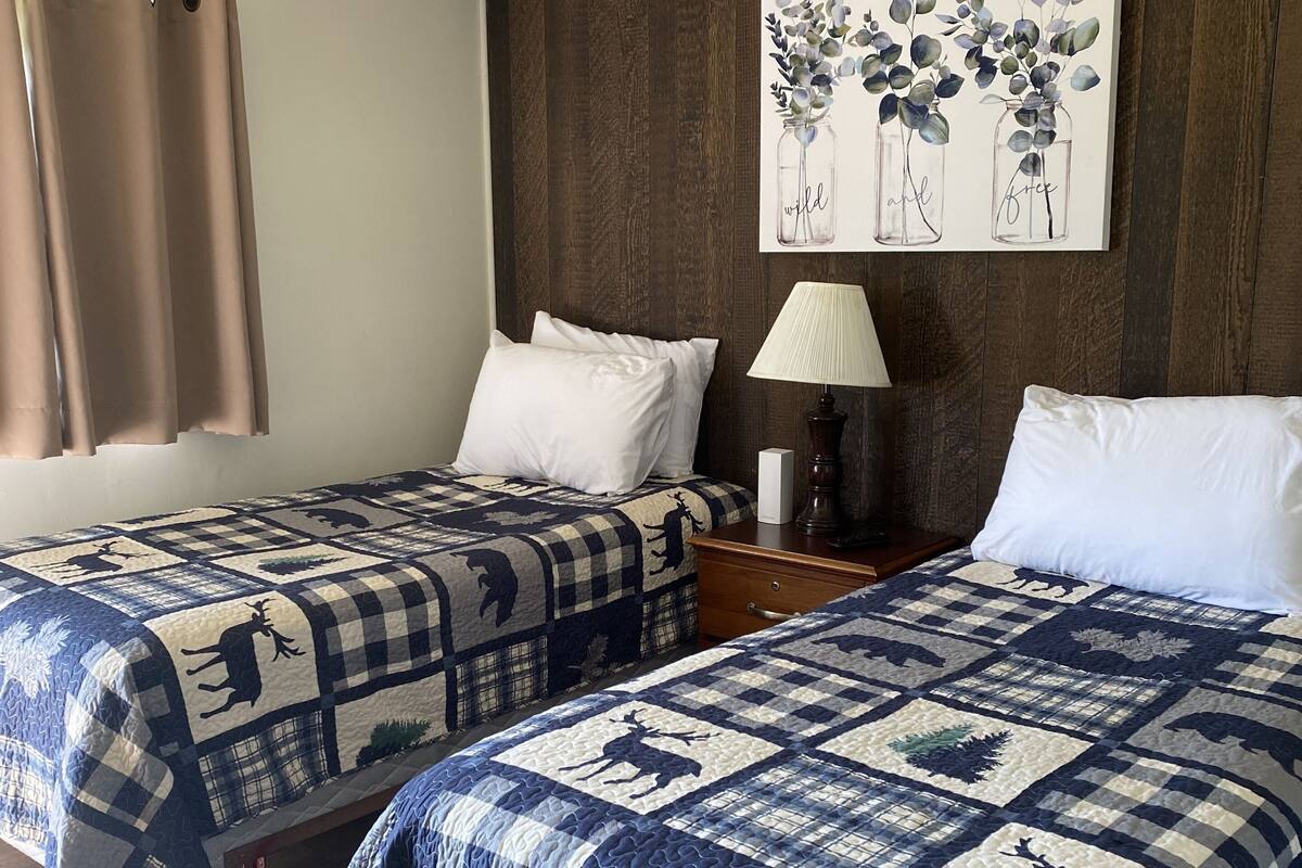 A hotel room with two beds, each with a blue and white checkered quilt featuring animal prints. There is a ceiling fan, a painting of eucalyptus leaves on the wall, and a lamp on a wooden nightstand.