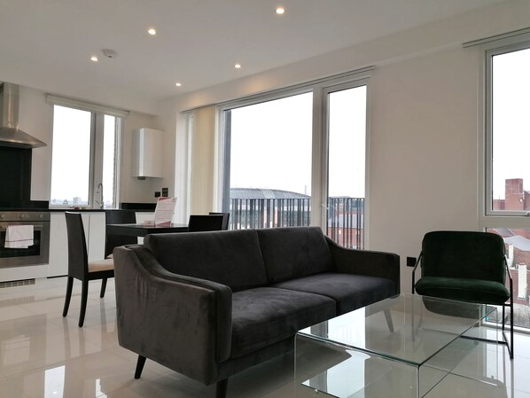 A modern living room with a glass coffee table, a dark grey sofa, and a green chair. There is a kitchen area with a stove and a dining table with chairs. Large windows provide a view of the cityscape. - Still Life Euston Deluxe (London)
