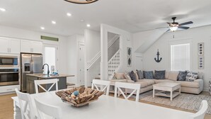 A modern kitchen with white cabinetry, a built-in oven, and a refrigerator. There is a dining area with a white table and chairs, and a living room with a beige sofa and a coffee table. - 30A - The Charming Blue Haven by PHG (Santa Rosa Beach)
