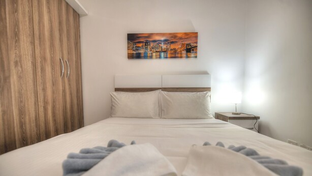 A neatly made bed with white linens and a blue towel, a wooden wardrobe, a nightstand with a lamp, and a cityscape painting on the wall. - Spacious & modern 3 bedroom near seafront JBOR1-1 (Gzira)