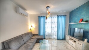 A living room with a grey sofa, a glass coffee table, a TV on a wooden stand, and blue curtains. - Spacious & modern 3 bedroom near seafront JBOR1-1 (Gzira)