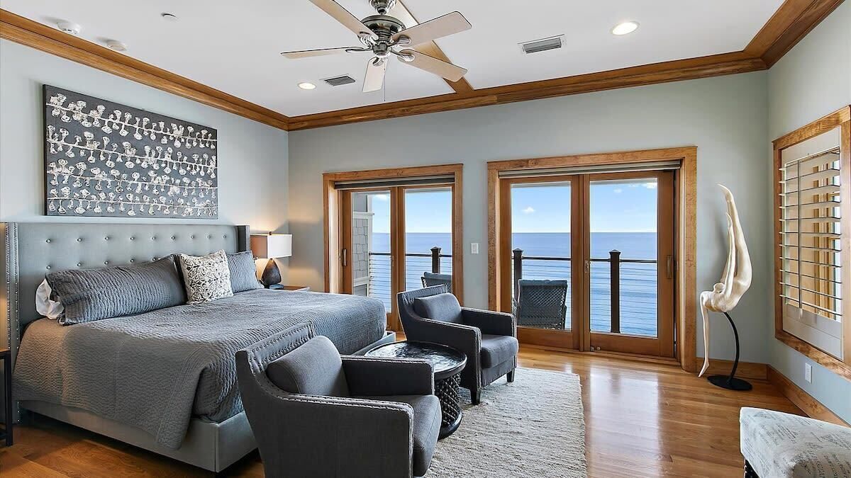 A bedroom with a large bed, two armchairs, a ceiling fan, and a view of the ocean through large windows.