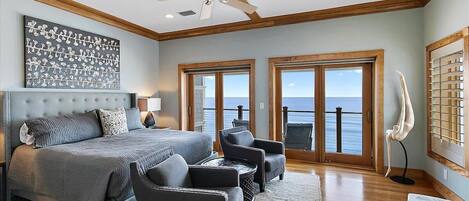 A bedroom with a large bed, two armchairs, a ceiling fan, and a view of the ocean through large windows.