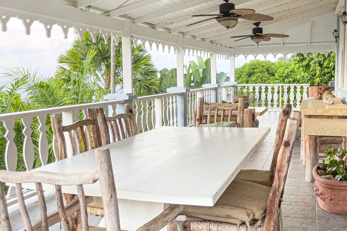A covered outdoor dining area with a white table and wooden chairs, surrounded by a white railing and potted plants.