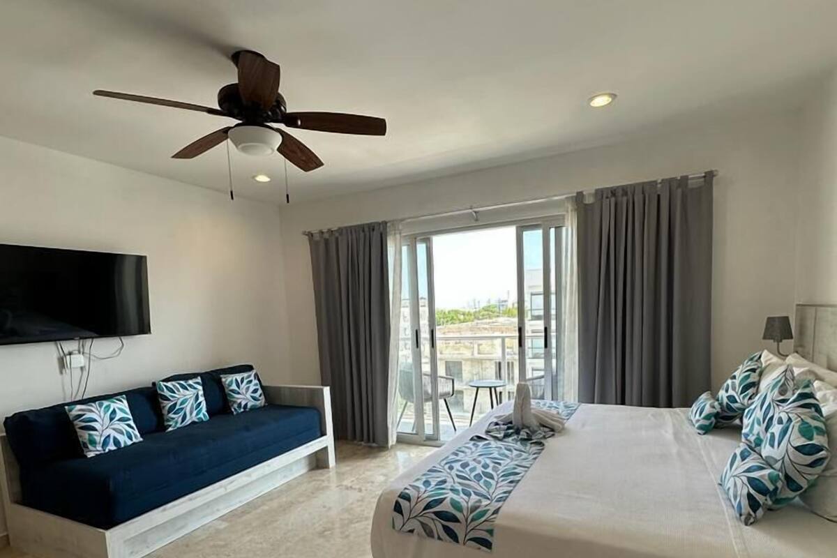 A hotel room with a bed, a sofa, a ceiling fan, a TV, and a balcony with curtains.