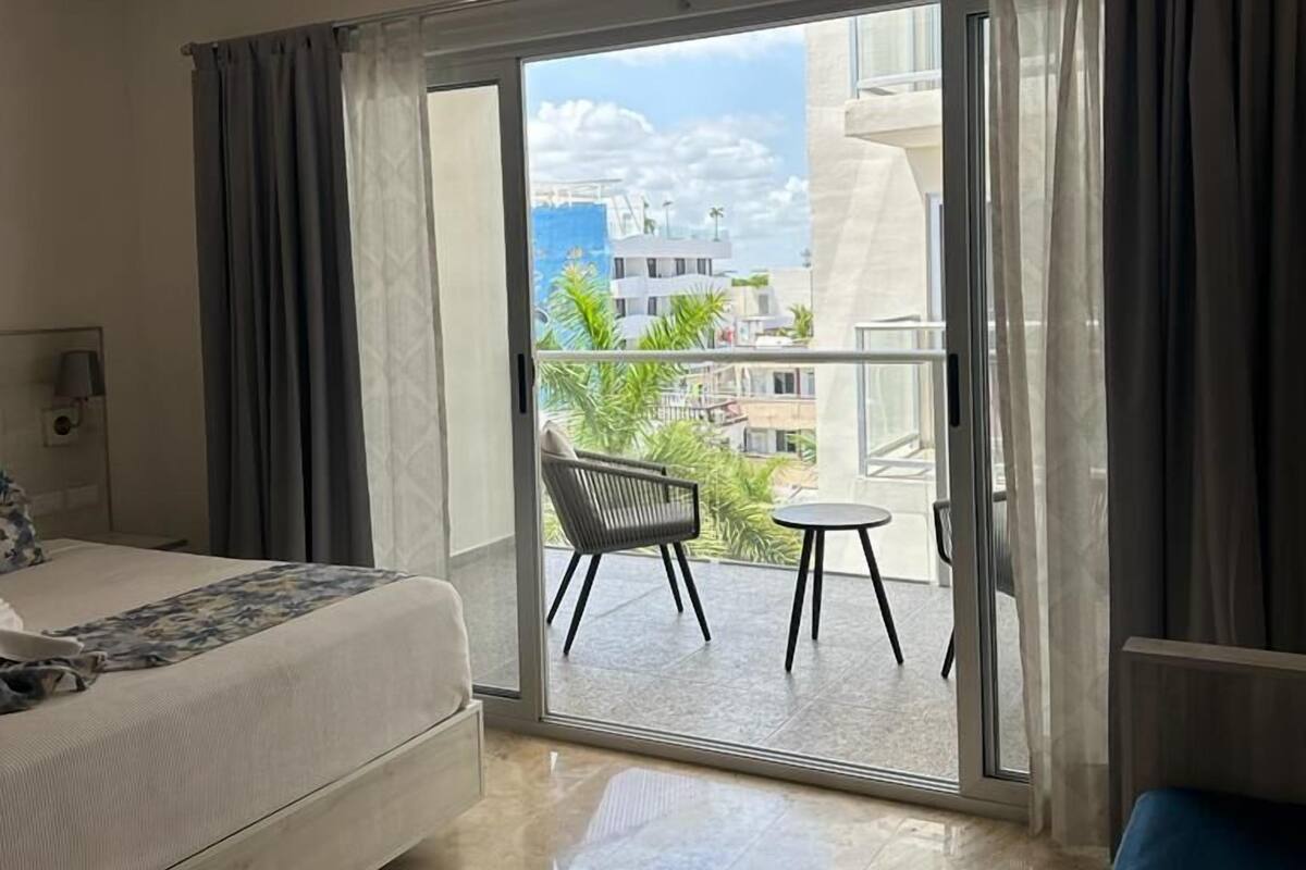 A hotel room with a bed, a small table, and a chair on a balcony with a city view.