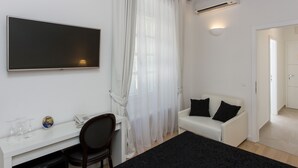 A modern room with a flat-screen TV, a desk with a chair, a white sofa with black cushions, and a large window with sheer curtains.