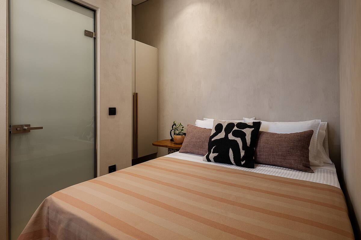 Un dormitorio con cama, almohadas, una mesita de noche y una planta en maceta.