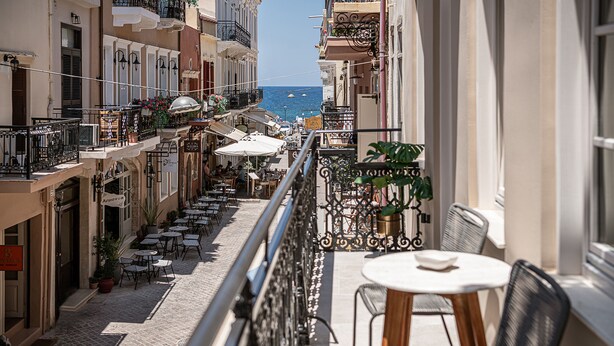 A balcony with a view of a coastal street, outdoor seating, and potted plants. - Casa Modiano (Chania)