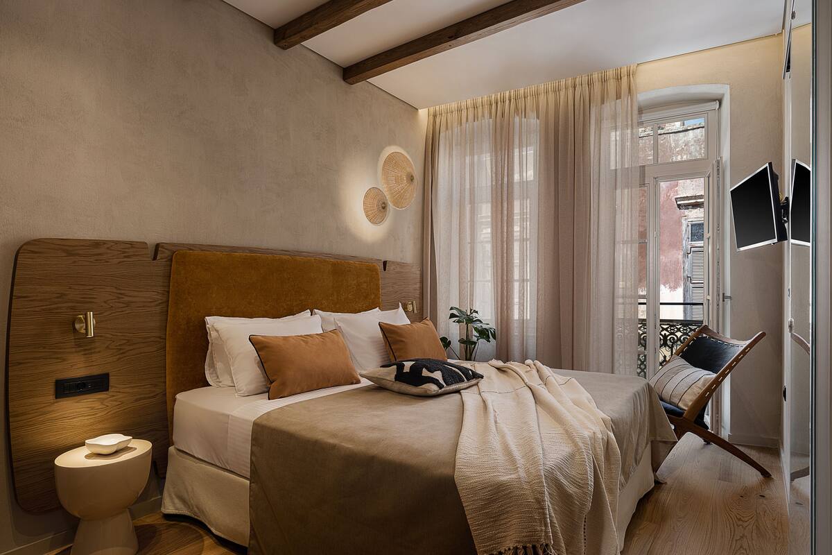 Un dormitorio con cabecera de madera, una cama con varios almohadones, una mesita de noche, un inodoro moderno y una ventana con cortinas.