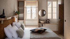 A bedroom with a bed, pillows, a bedside table with a plant, a mirror, and a window with a view. - Casa Modiano (Chania)