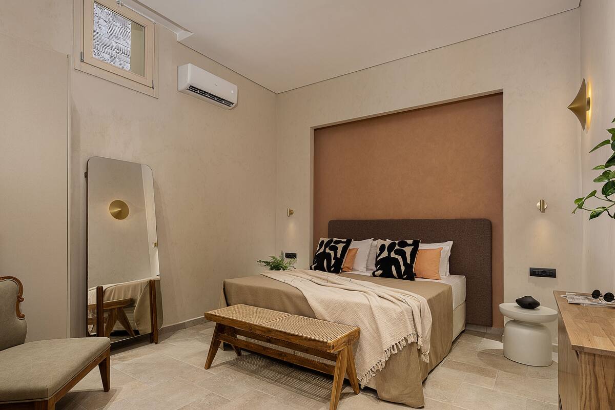 Un dormitorio moderno con una cama grande, una mesita de noche, un banco, un espejo y un equipo de aire acondicionado.