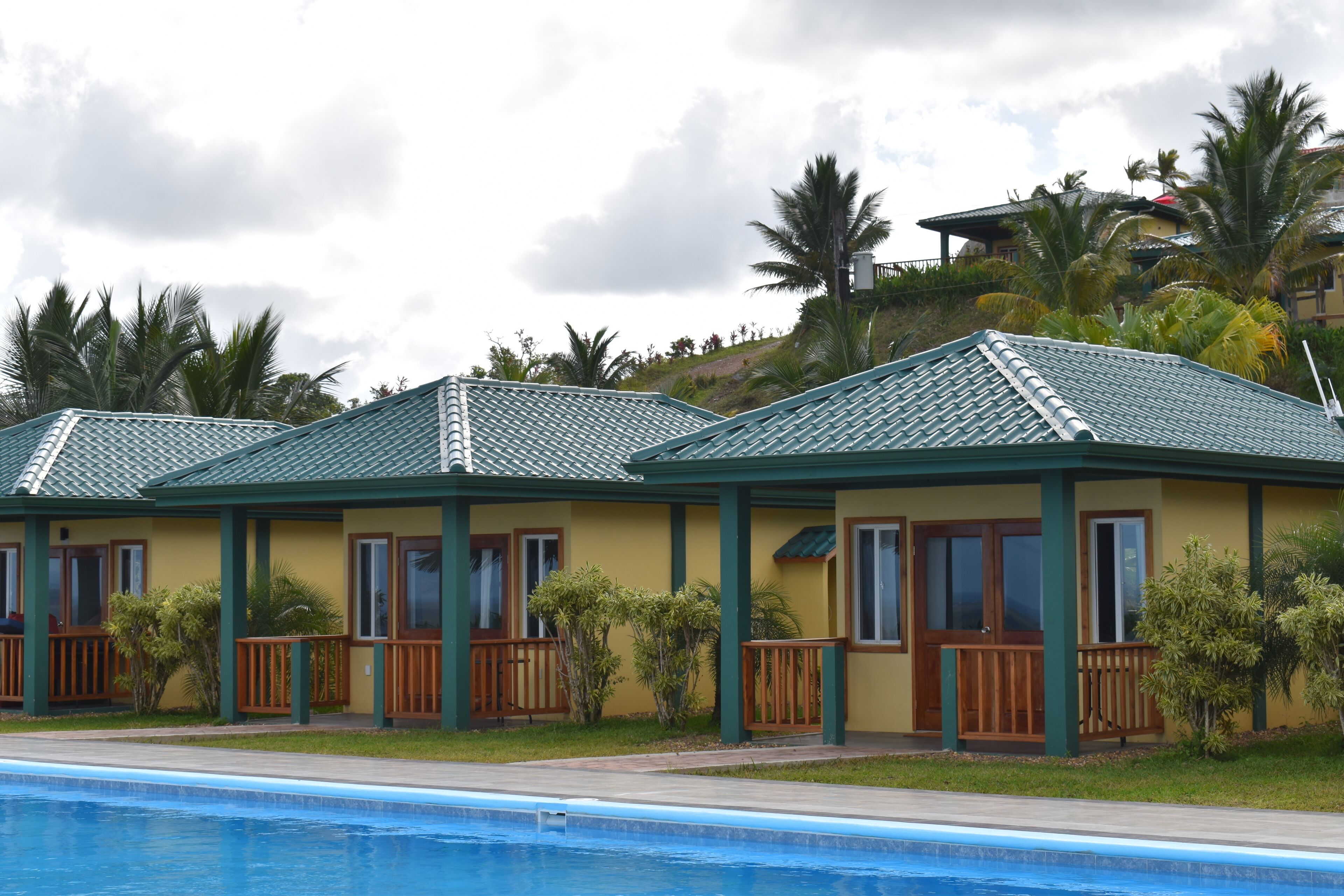 A row of small, single-story buildings with green roofs and wooden railings, adjacent to a swimming pool.