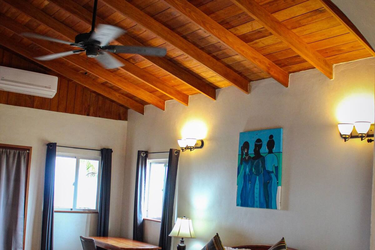 A bedroom with a wooden ceiling, a bed with pillows, a desk, and a painting on the wall.