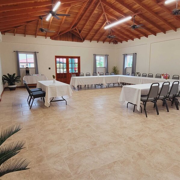 Une salle de conférence spacieuse, dotée de hauts plafonds et de poutres en bois, équipée de ventilateurs de plafond et baignée de lumière naturelle grâce à de grandes fenêtres.