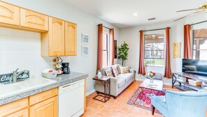 A modern kitchen with wooden cabinets, a dishwasher, and a coffee station. There is a living area with a sofa, coffee table, and a TV. - Arizona Desert Vacation Rental w/ Pool Access! (Eloy)