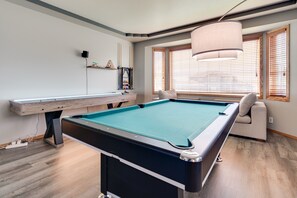 Una mesa de billar en una habitación con piso de madera, un banco, un sofá y una ventana con persianas.