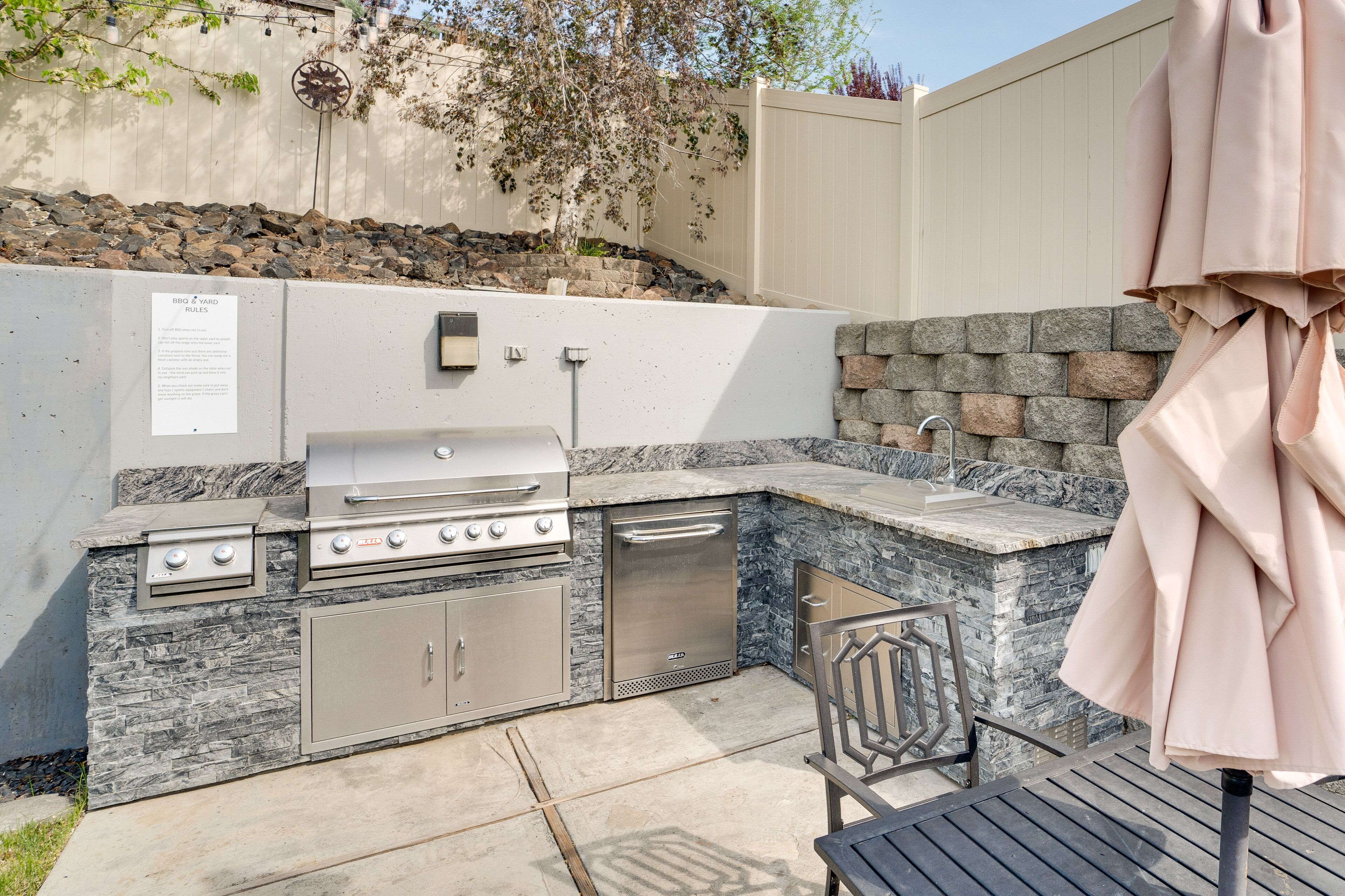 Cucina esterna con barbecue, lavello e pensili, affacciata su un muro di pietra e un giardino roccioso.