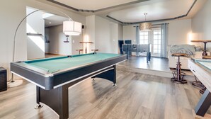 Una mesa de billar en una sala amplia con suelo de madera, un diseño de techo moderno y una zona de estar con sofá y mesa.