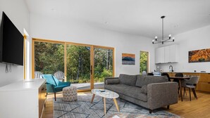 A modern living room with a sofa, coffee table, and chairs, a kitchen area with cabinets and a dining table, and large sliding glass doors to a deck with a view of trees. - KOTI 1 (Two Bedroom) Traverse City's Scandinavian Cabins, vacation home on TART & mins to Traverse C... (Williamsburg)