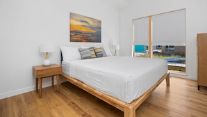 Una camera da letto con un letto in legno, biancheria bianca, un comodino con una lampada, un dipinto di un tramonto e un’ampia finestra con tende.