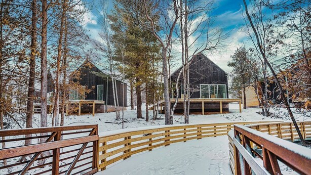 Eine Holzterrasse führt zu einer Hütte mit Glasfassade, umgeben von Bäumen und Schnee.