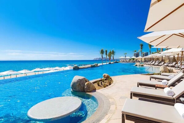 Relax by the ocean with a pool, hot tub, and waterfall, surrounded by lounge chairs and umbrellas.
