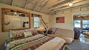 A rustic room with a bed, a wooden ceiling, a large mirror, an American flag, and a window with blinds. - Cozy Lakefront Cabin w/ Sandy Beach & Bunkhouse! (Deerwood)