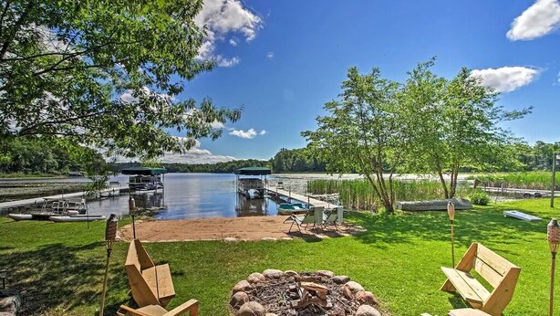 A lakeside patio with a fire pit, wooden chairs, and a dock with boats. - Cozy Lakefront Cabin w/ Sandy Beach & Bunkhouse! (Deerwood)