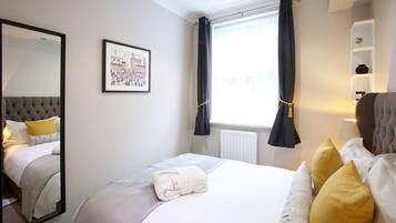 Quarto de hotel com cama, um espelho grande, janela com cortinas, radiador e prateleira com uma planta.