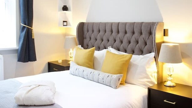 A neatly made bed with a grey headboard, white bedding, and a few pillows. There are two bedside tables with lamps, a wall-mounted shelf with decorative items, and a window with a curtain. - Avari - Luxury 2BR Apartment • Steps from Victoria (London)