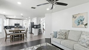 A modern kitchen with white cabinets, a dining area with a wooden table and chairs, a living room with a grey sofa and patterned cushions, and a ceiling fan.