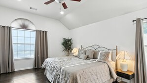 A bedroom with a large bed, two bedside lamps, a ceiling fan, and a window with curtains.