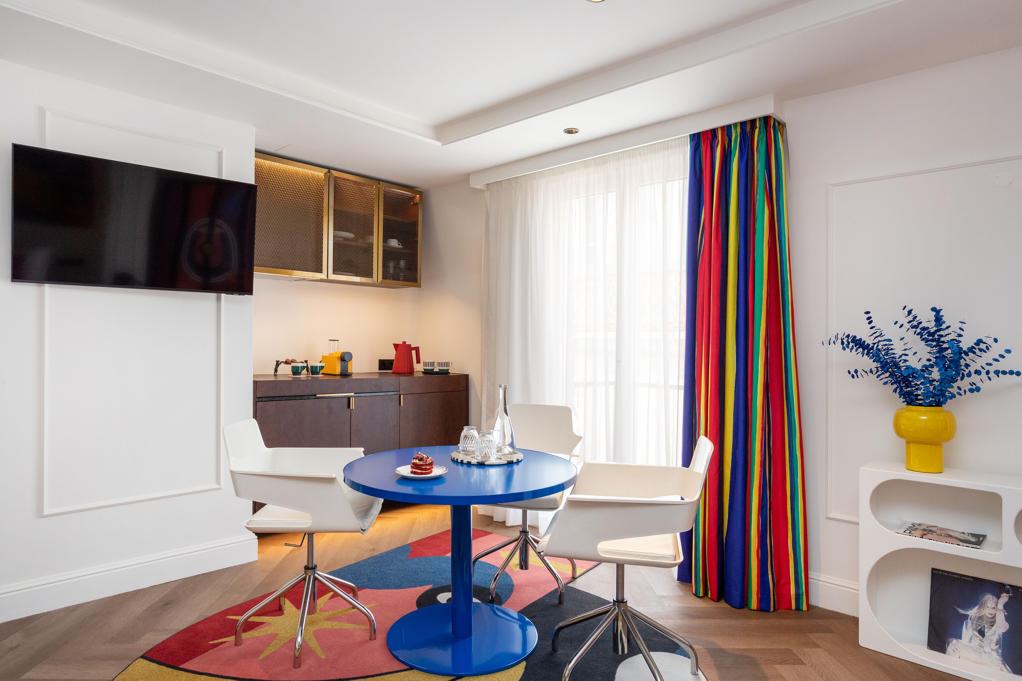Un salon moderne avec une table ronde bleue, des chaises blanches, un téléviseur à écran plat fixé au mur, un coin cuisine avec un meuble en bois et un rideau rayé aux couleurs vives.
