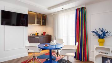 Un salon moderne avec une table ronde bleue, des chaises blanches, un téléviseur à écran plat fixé au mur, un coin cuisine avec un meuble en bois et un rideau rayé aux couleurs vives.