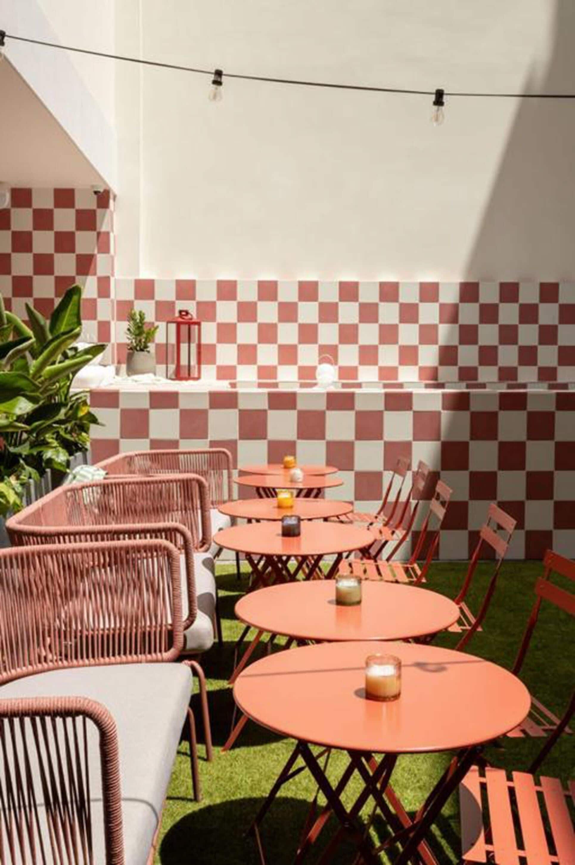 An outdoor patio with round tables and wicker chairs, a checkered wall, and string lights.