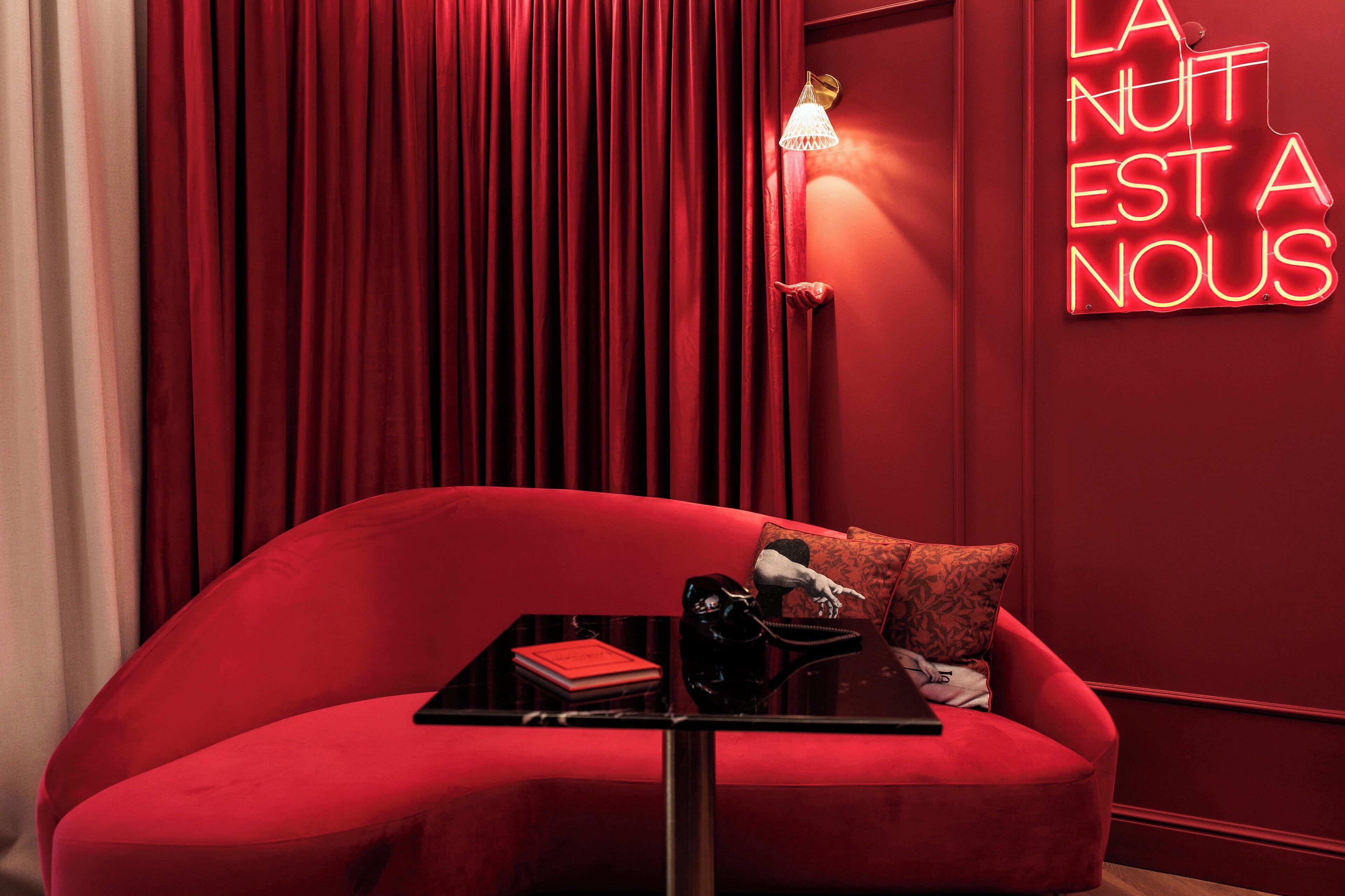 Un canapé rouge avec une petite table noire et un livre rouge, un sac à main noir et une lampe, le tout placé devant un mur rouge orné d'un néon.