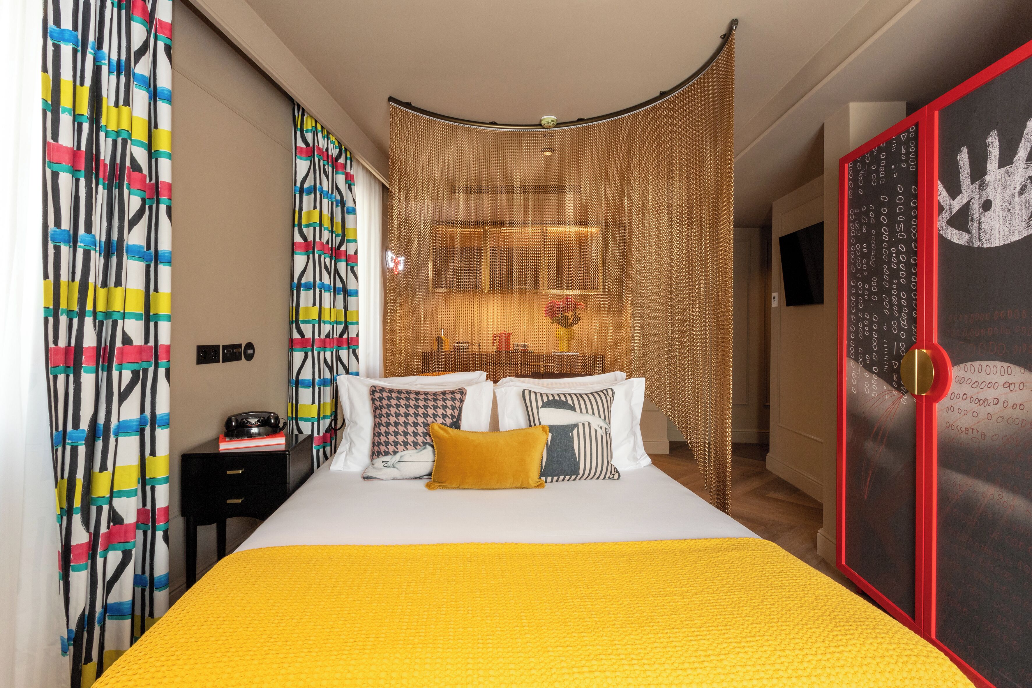 Une chambre d’hôtel moderne avec un grand lit, des rideaux colorés, un paravent en osier et une œuvre d’art encadrée de rouge accrochée au mur.