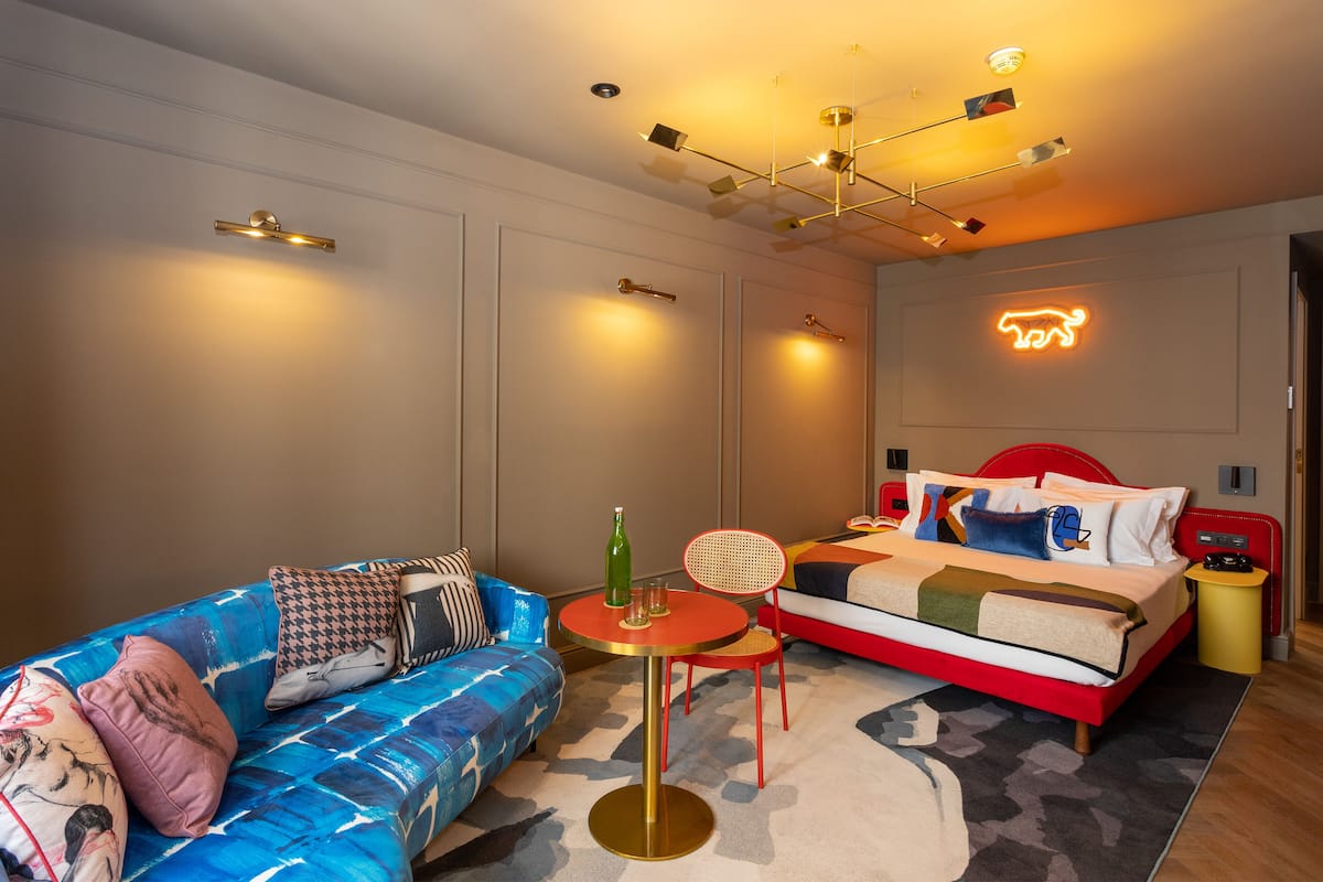 Moderni hotellihuone, jossa on sininen sohva, pyöreä pöytä pullolla, punainen sänky ja seinällä loistava neonvalomainos, jossa on eläinaihe.