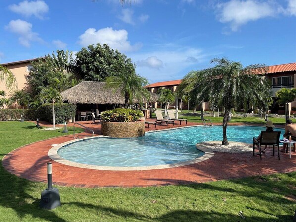A swimming pool with a brick border, surrounded by grass and palm trees. - Amazing 2-Bedroom Townhouse near Eagle Beach (Eagle Beach)