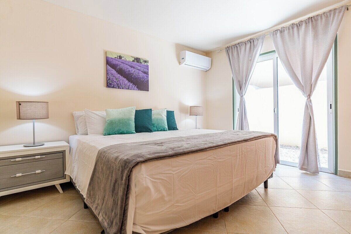 Un dormitorio con cama, mesitas de noche, lámpara, aire acondicionado y ventana con cortinas.